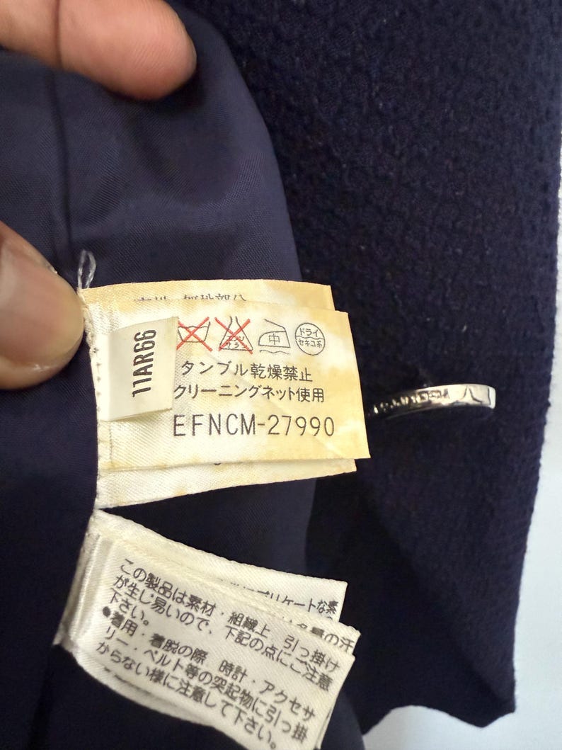 May include: Close-up of a navy blue garment with a tag featuring Japanese text and care symbols. The tag includes the code "11AR66" and "EFNCM-27990". The garment has a silver-colored clasp.