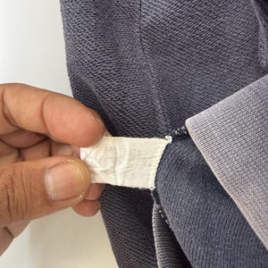 May include: Close-up of a section of a dark blue garment, possibly a sweatshirt or hoodie. The fabric has a textured appearance. A white tag with text is held in the frame, near the garment's seam.