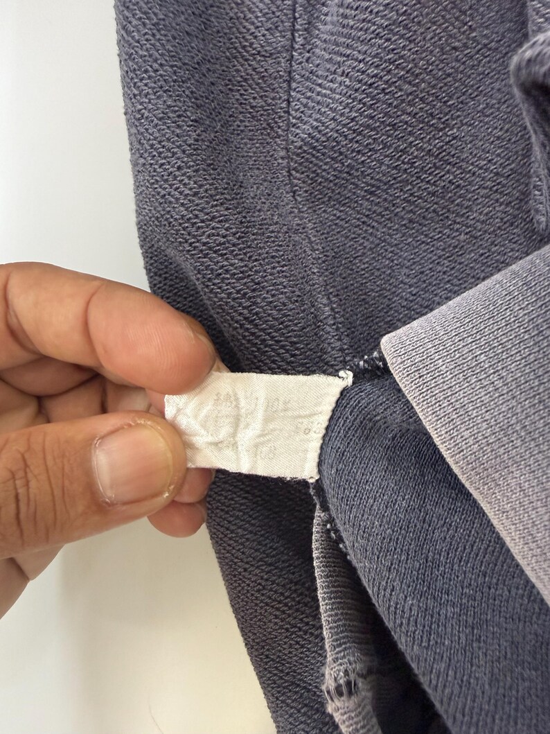 May include: Close-up of a section of a dark blue garment, possibly a sweatshirt or hoodie. The fabric has a textured appearance. A white tag with text is held in the frame, near the garment's seam.