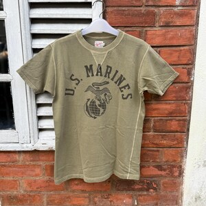 May include: An olive green short-sleeve t-shirt featuring a dark brown graphic print on the chest. The print displays the text U.S. MARINES arched over an eagle, globe, and anchor emblem with the banner text SEMPER FIDELIS. The shirt has a crew neck.