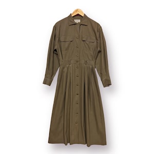 Peut inclure: Robe boutonnée vert olive à manches longues, deux poches poitrine et une taille ceinturée. La robe est suspendue à un cintre en bois. La robe a un col chemise et un ourlet au genou.