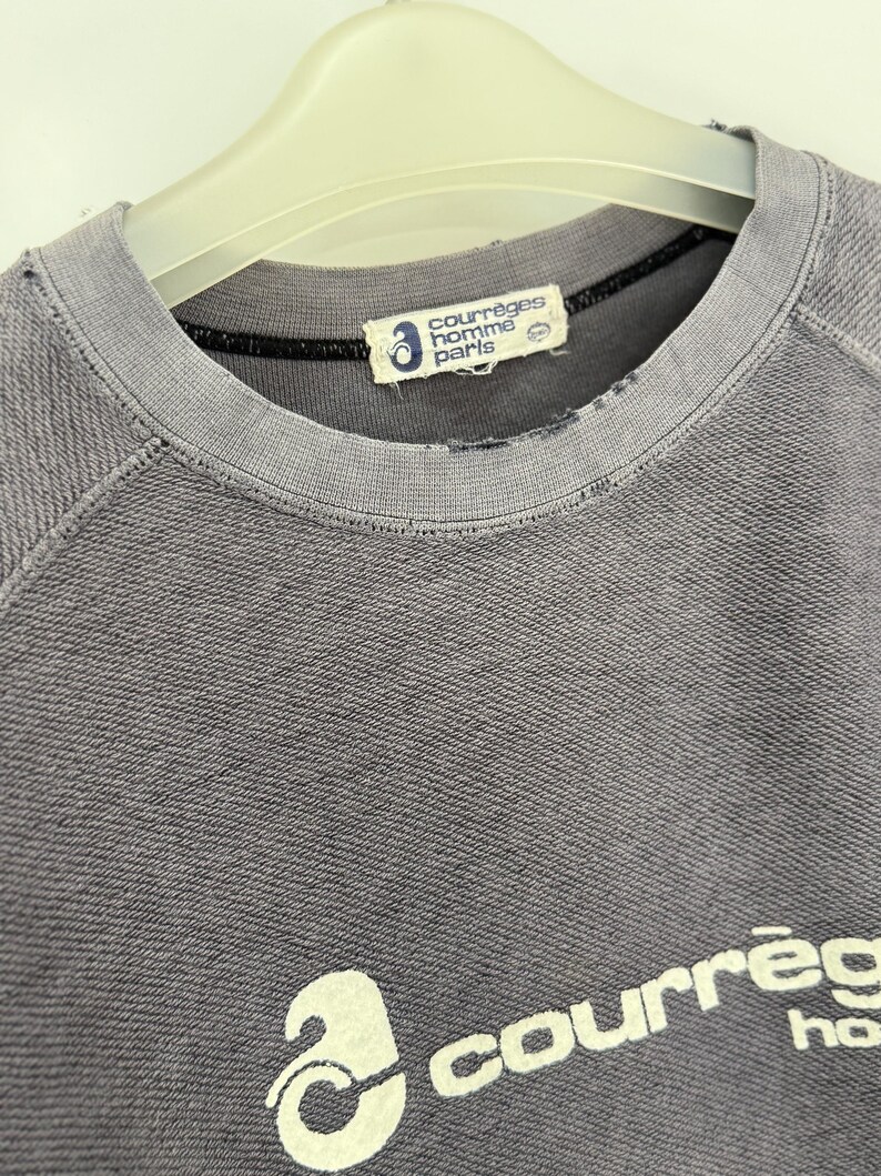 May include: A gray crew neck sweatshirt with the brand name "courr&egrave;ges homme paris" printed on the front. The sweatshirt has a distressed look, with a worn collar and a faded logo.