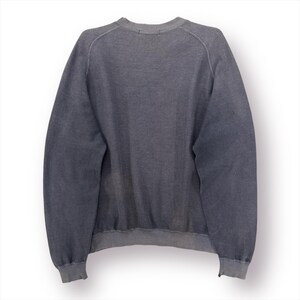 May include: A faded gray sweatshirt with a crew neck and long sleeves. The garment has ribbed cuffs and hem. The fabric appears to be a soft, worn material, suggesting comfort and casual wear. The back view of the sweatshirt is shown.