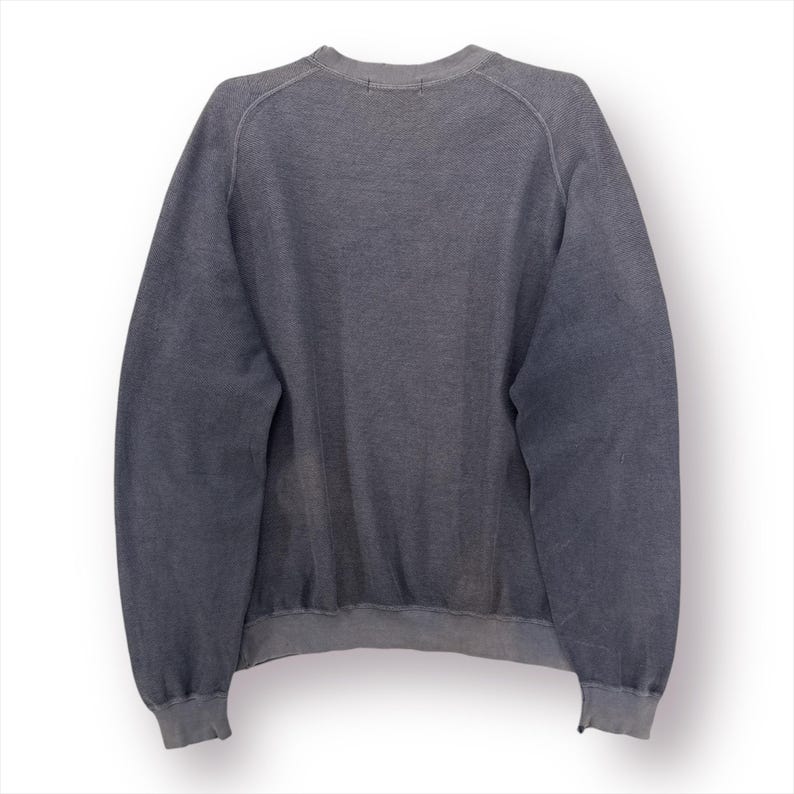 May include: A faded gray sweatshirt with a crew neck and long sleeves. The garment has ribbed cuffs and hem. The fabric appears to be a soft, worn material, suggesting comfort and casual wear. The back view of the sweatshirt is shown.