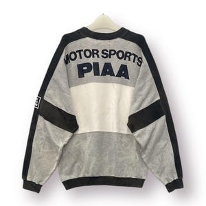 May include: A gray, white, and black sweatshirt with the text "MOTOR SPORTS PIAA" across the back. The sweatshirt has a color block design with black accents on the sleeves and shoulders. The garment is hanging on a white hanger.