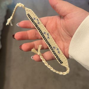 May include: A handmade friendship bracelet in cream and black, featuring a woven design with small blue accents. The bracelet has a braided cord closure and is held in a hand.