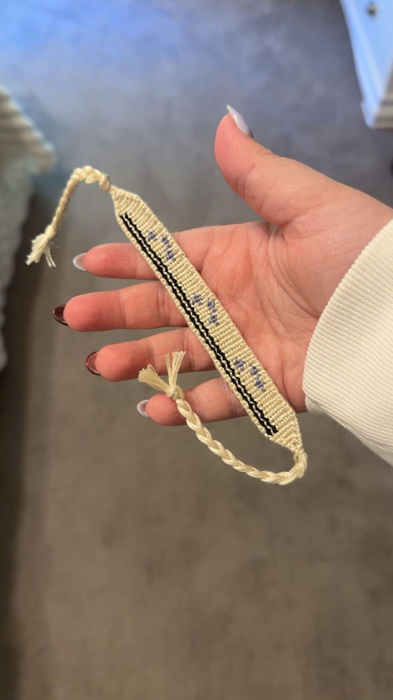 May include: A handmade friendship bracelet in cream and black, featuring a woven design with small blue accents. The bracelet has a braided cord closure and is held in a hand.