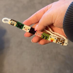 以下が含まれることがあります： カラフルなピクセル化された顔のパターンが施された白い友情ブレスレット。ブレスレットは刺繍糸で作られており、シンプルな結び目の留め具が付いています。