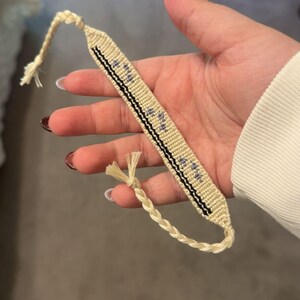 May include: A handmade friendship bracelet in cream and black, featuring a woven design with small blue accents. The bracelet has a braided cord closure and is held in a hand.