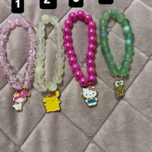 May include: Four beaded bracelets with cartoon character charms. The bracelets are in pink, white, and green. Each bracelet has a different charm: My Melody, Pikachu, Hello Kitty, and Keroppi.