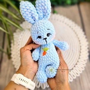Puede incluir: Un juguete de conejo de crochet azul claro con una peque&ntilde;a zanahoria bordada en el pecho. El conejo est&aacute; siendo sostenido por las manos de una persona.