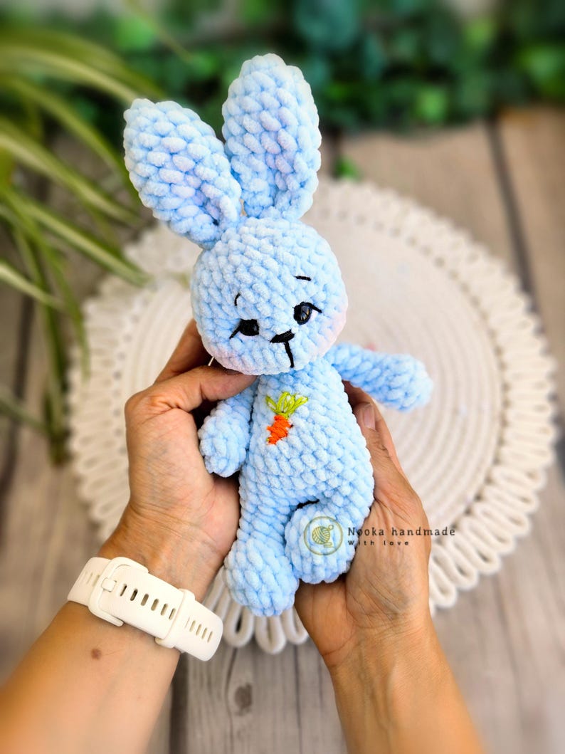 Puede incluir: Un juguete de conejo de crochet azul claro con una peque&ntilde;a zanahoria bordada en el pecho. El conejo est&aacute; siendo sostenido por las manos de una persona.