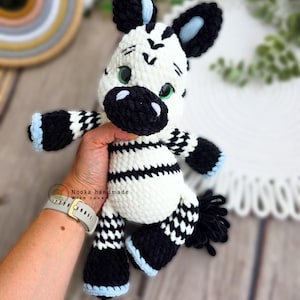 Può includere: Un peluche a forma di zebra fatto a mano all'uncinetto. La zebra ha un corpo bianco con strisce nere, zampe a scacchi bianchi e neri e orecchie nere con accenti azzurri. Il giocattolo ha occhi verdi e una coda nera.
