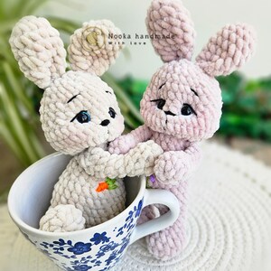Puede incluir: Dos juguetes de peluche de conejo de crochet, uno rosa y otro beige, est&aacute;n sentados en una taza de t&eacute; azul y blanca. El conejo rosa est&aacute; sosteniendo la mano del conejo beige. El texto "Nooka handmade with love" es visible en el fondo.