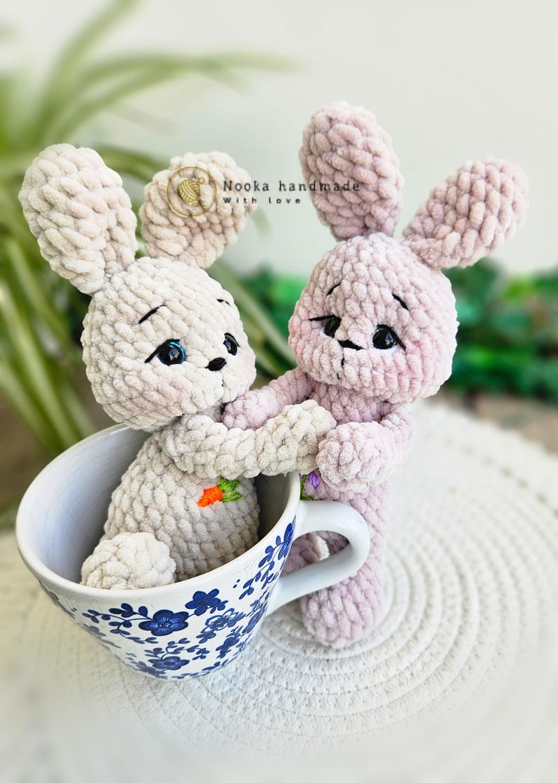 Puede incluir: Dos juguetes de peluche de conejo de crochet, uno rosa y otro beige, est&aacute;n sentados en una taza de t&eacute; azul y blanca. El conejo rosa est&aacute; sosteniendo la mano del conejo beige. El texto "Nooka handmade with love" es visible en el fondo.