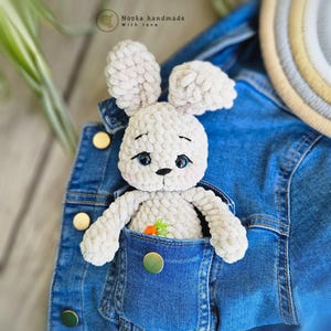 Puede incluir: Un conejito de crochet blanco con ojos azules y una zanahoria bordada en el pecho est&aacute; sentado en el bolsillo de una chaqueta vaquera azul. El texto "Nooka handmade with love" es visible sobre el conejito.