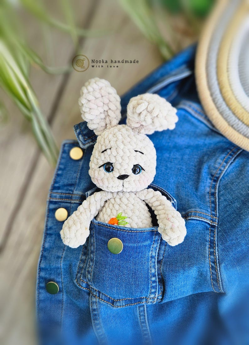 Puede incluir: Un conejito de crochet blanco con ojos azules y una zanahoria bordada en el pecho est&aacute; sentado en el bolsillo de una chaqueta vaquera azul. El texto "Nooka handmade with love" es visible sobre el conejito.