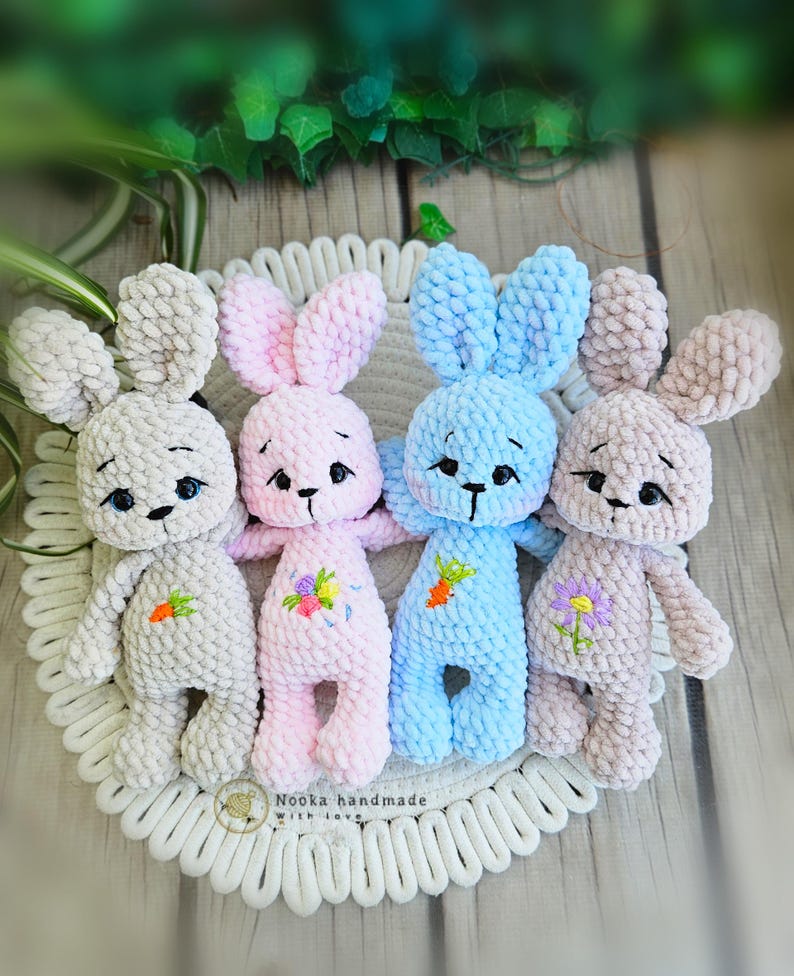 Puede incluir: Cuatro peluches de conejo de crochet en diferentes colores: gris, rosa, azul y beige. Cada conejo tiene detalles bordados, incluidos ojos, nariz y boca. Los conejos est&aacute;n sentados en una alfombra de crochet blanca.