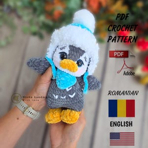 Op de afbeelding: Een handgemaakte gehaakte pinguïnpop met een wit-blauwe hoed, blauwe sjaal en gele voeten. De pinguïn is grijs en wit met een gele snavel. De afbeelding bevat de tekst "PDF Crochet Pattern" en vlaggen voor Roemenië en de Verenigde Staten.