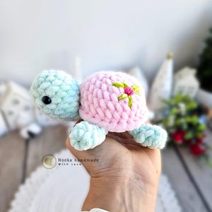 Può includere: Un giocattolo di tartaruga all'uncinetto con un guscio rosa e un corpo blu chiaro. Il guscio ha un fiore verde ricamato su di esso. La tartaruga è tenuta in una mano.