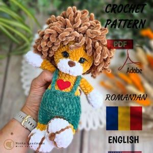 Può includere: Una bambola leone all'uncinetto fatta a mano con una criniera marrone, corpo giallo e salopette verde. La bambola ha zampe bianche e un cuore rosso sul petto. L'immagine include il testo "PDF Crochet Pattern" e "English".
