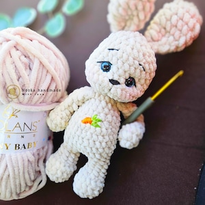 Puede incluir: Un conejito de peluche blanco tejido a crochet con ojos bordados y una zanahoria en el pecho. El conejito est&aacute; sentado sobre una superficie marr&oacute;n junto a una bola de hilo rosa con la etiqueta "Nooka handmade with love".