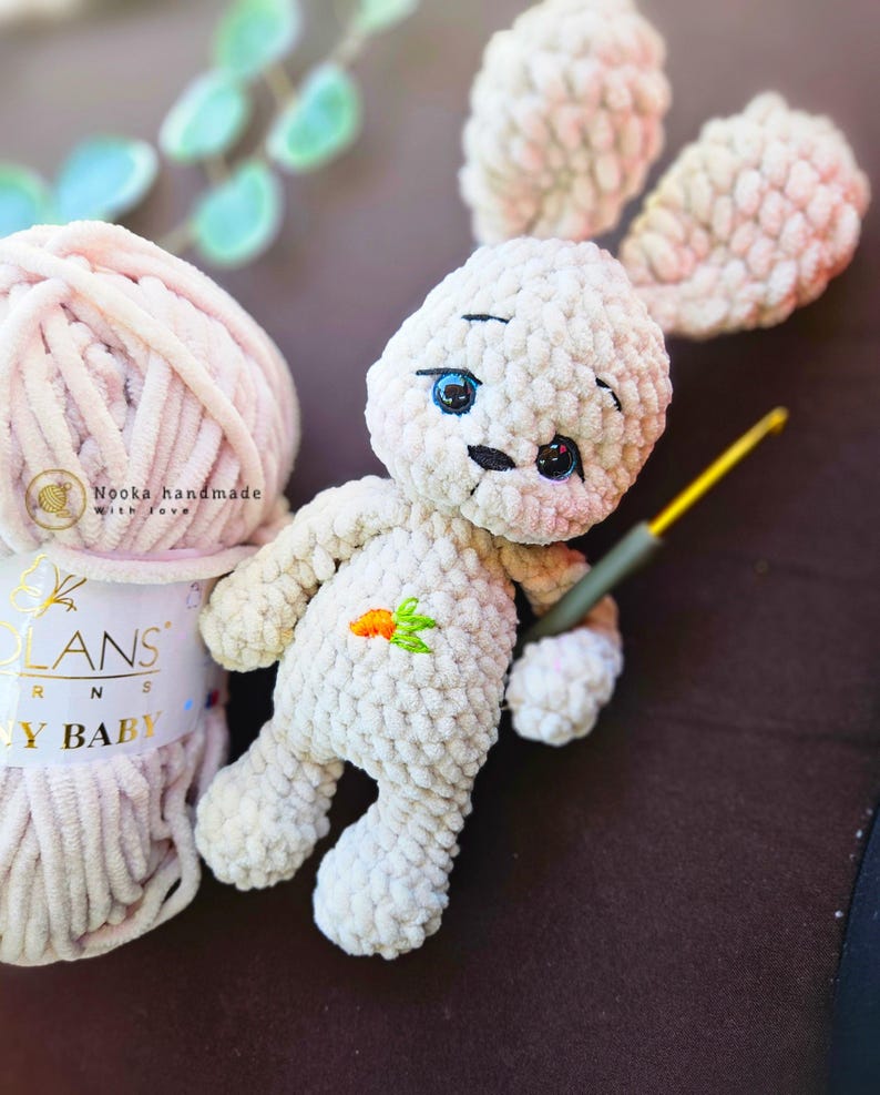 Puede incluir: Un conejito de peluche blanco tejido a crochet con ojos bordados y una zanahoria en el pecho. El conejito est&aacute; sentado sobre una superficie marr&oacute;n junto a una bola de hilo rosa con la etiqueta "Nooka handmade with love".