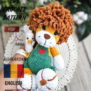 Pode incluir: Padrão de crochê PDF para um brinquedo amigurumi de leão com juba marrom, macacão verde e um pintinho branco. O padrão está disponível em inglês e romeno. O leão está sentado em um tapete de crochê branco.