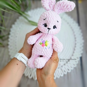 Puede incluir: Un conejito de crochet rosa con flores bordadas en el pecho. El conejito tiene ojos negros y una nariz negra. El conejito est&aacute; siendo sostenido por la mano de una persona.