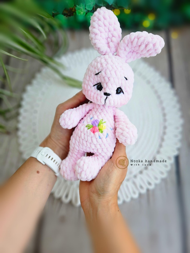 Puede incluir: Un conejito de crochet rosa con flores bordadas en el pecho. El conejito tiene ojos negros y una nariz negra. El conejito est&aacute; siendo sostenido por la mano de una persona.