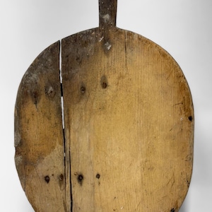 May include: A round, wooden cutting board with a handle. The board is made of light-colored wood with visible grain and nail heads. It has a rustic, worn appearance, with a crack running down the center. The handle is rectangular and attached to the top.