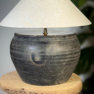 May include: A black ceramic table lamp with a cream-colored lampshade. The lamp has a round, textured base and a brass-colored stem. The lamp is sitting on a small, round wooden stool.