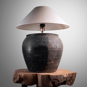 May include: A rustic lamp with a dark, textured ceramic base and a light-colored, conical lampshade. The lamp sits on a natural wood side table, showcasing a blend of textures and tones. The lamp's design suggests a vintage or antique aesthetic.