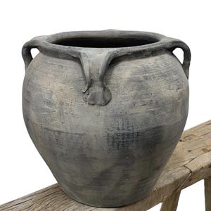 May include: A large, gray ceramic pot with two handles and a textured surface. The pot has a wide opening and a rounded body. It is sitting on a wooden bench. The pot is suitable for indoor or outdoor use.