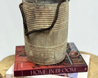 May include: A weathered, cylindrical wooden bucket with horizontal grooves and a dark brown rope handle. The bucket sits atop two books: "Home in Bloom" and "Livable Luxe". The books are on a light-colored, wooden surface.
