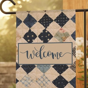 May include: A garden flag with a quilted pattern in shades of blue and white, featuring the word "welcome" in a cursive font. The flag is displayed on a black metal stand, with a backdrop of greenery and flowers.