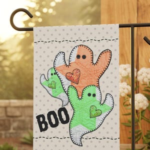 May include: A rectangular garden flag featuring a light gray background with white polka dots. The design displays three ghost figures in orange, light green, and white, each with a small heart and black stitching. The word BOO is printed in black.
