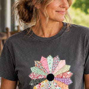 May include: A charcoal gray short-sleeve t-shirt featuring a large, circular, multi-colored floral applique on the chest. The flower is composed of various patterned fabric segments in shades of pink, green, red, and white, with a solid black center.