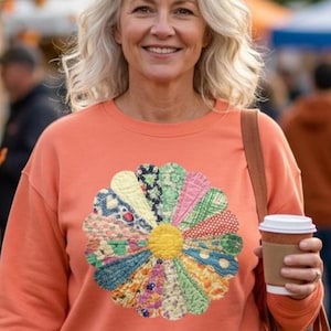 May include: A peach-colored crewneck sweatshirt featuring a large, circular, multi-colored floral applique on the chest. The flower is composed of various patterned fabric petals in shades of green, yellow, red, and blue, with a yellow center.