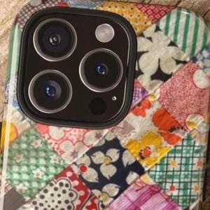 May include: A smartphone case featuring a colorful patchwork design with various floral, striped, and checkered patterns in shades of green, pink, yellow, and blue. The case has a large black camera cutout at the top with three circular lenses.