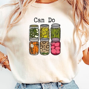 May include: A white t-shirt with the text "Can Do" printed on the front. The text is above a graphic of six glass jars filled with different types of preserved vegetables.