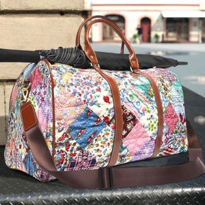 May include: A patterned travel bag with brown leather handles and straps. The bag is made of patchwork fabric with floral and geometric designs in blue, pink, and green. A black umbrella is placed on top.