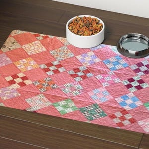 May include: A rectangular pet feeding mat featuring a patchwork quilt pattern in shades of pink, coral, and white with small squares of blue, green, and red. The mat is positioned under a white ceramic bowl filled with dry food and a metal bowl.