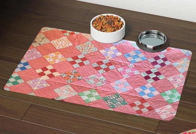 May include: A rectangular pet feeding mat featuring a patchwork quilt pattern in shades of pink, coral, and white with small squares of blue, green, and red. The mat is positioned under a white ceramic bowl filled with dry food and a metal bowl.
