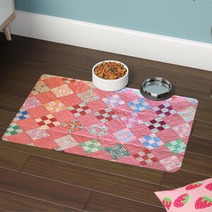 May include: A pink pet food mat with a patchwork quilt design. The mat features a variety of colorful squares in shades of pink, red, blue, and green. A white bowl filled with pet food and a silver bowl are on the mat.