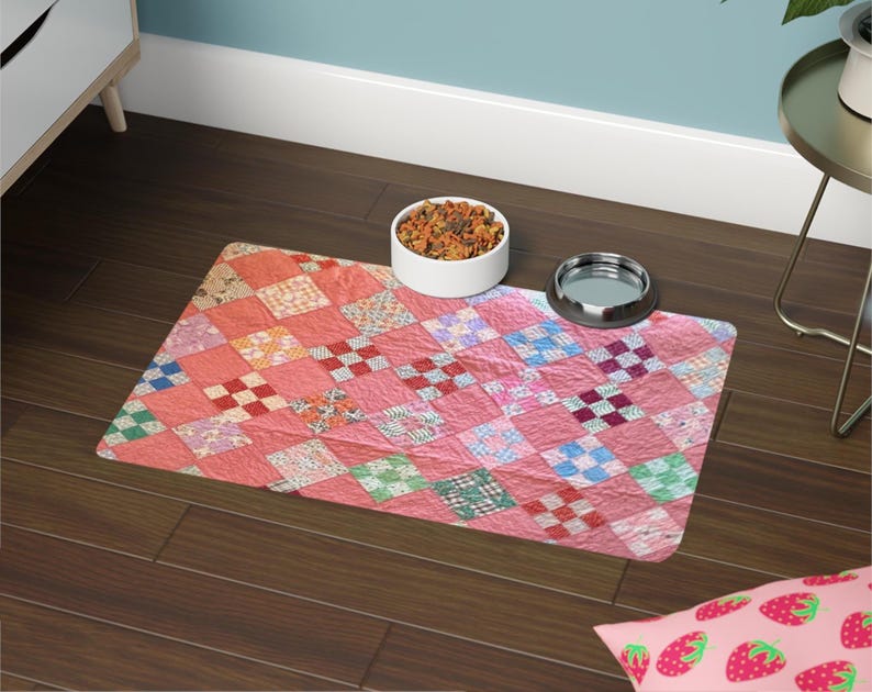 May include: A pink pet food mat with a patchwork quilt design. The mat features a variety of colorful squares in shades of pink, red, blue, and green. A white bowl filled with pet food and a silver bowl are on the mat.