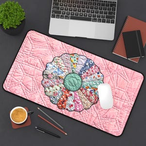 May include: A pink mousepad with a quilted pattern and a floral design in the center. The floral design is made up of various fabric patterns and colors. A white computer mouse sits on the right side.