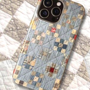 Puede incluir: Una funda para smartphone con un patr&oacute;n de tela acolchada en azul claro y una cuadr&iacute;cula de peque&ntilde;os cuadrados en tonos azul, crema y rojo. El dise&ntilde;o imita un edred&oacute;n de patchwork tradicional con l&iacute;neas de costura diagonales visibles.