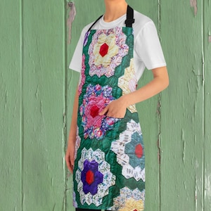May include: An apron with a colorful floral quilt pattern. The apron features a green background with hexagon-shaped floral designs in shades of red, pink, purple, and white. It has a black neck strap and a pocket.
