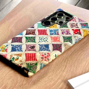 May include: A rectangular smartphone case featuring a multi-colored patchwork quilt pattern with various floral, checkered, and geometric prints separated by cream-colored stitched lines. The case has a black camera cutout area at the top.
